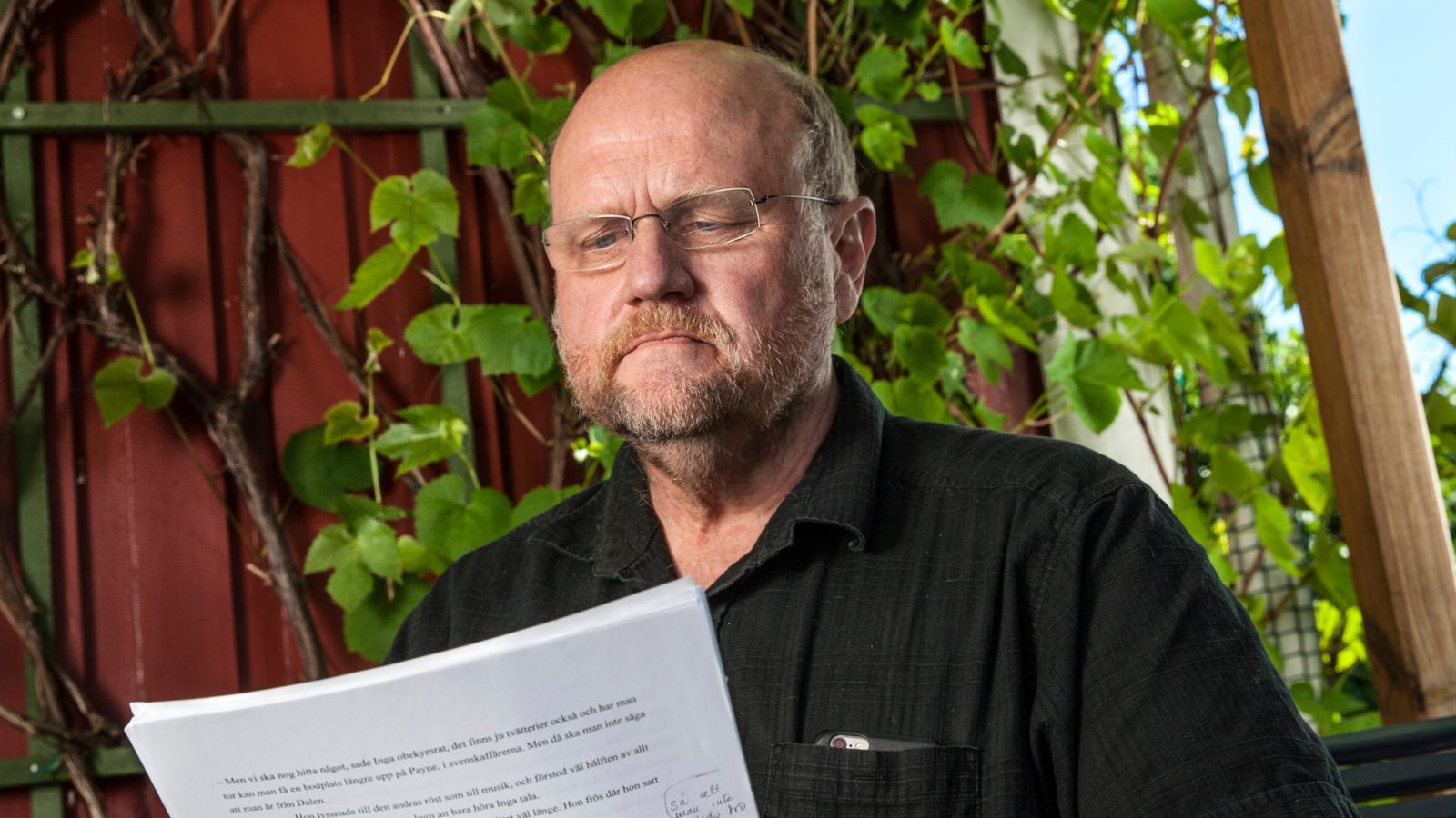 Ola Larsmo sitter i en trädgård och läser manuskript. Han är aktuell med boken Swede Hollow.