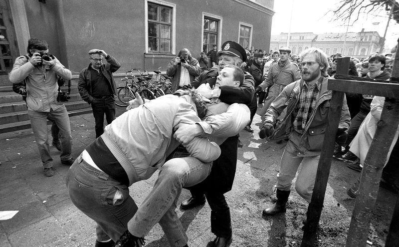 Bråk där en polis försöker sära två demonstranter. Folk i ring omkring och två fotografer bakom.