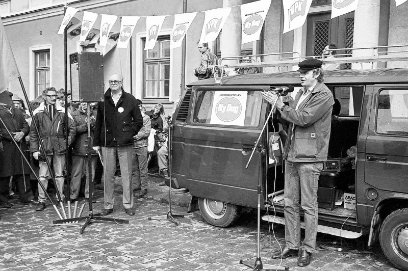 Lennart Värmby står framför en öppen Volkswagenbuss och talar i en mikrofon. Ovan hänger vimplar med VPK och arbetarpartitidningen Ny dag. Till vänster står Lars Werner.