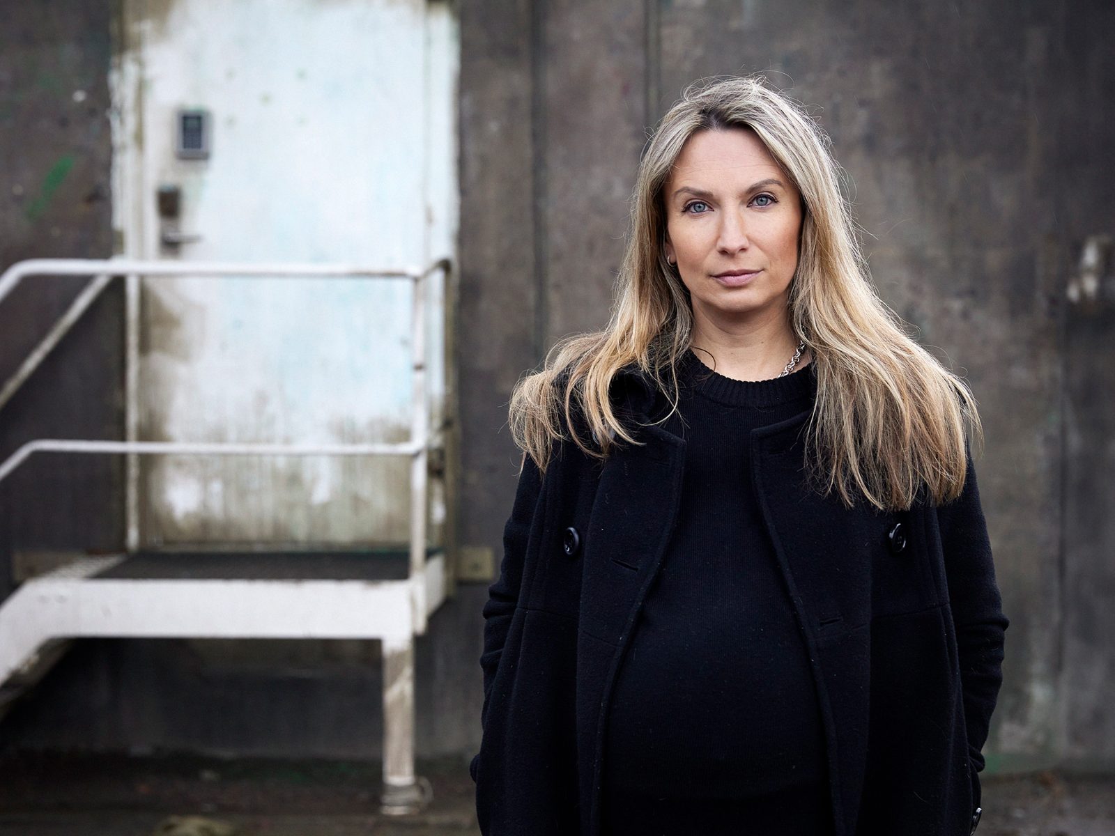 Porträtt på Valeria Helander framför en ruffig byggnad med en dörr med lås för att symbolisera kritiken mot sis-hem.