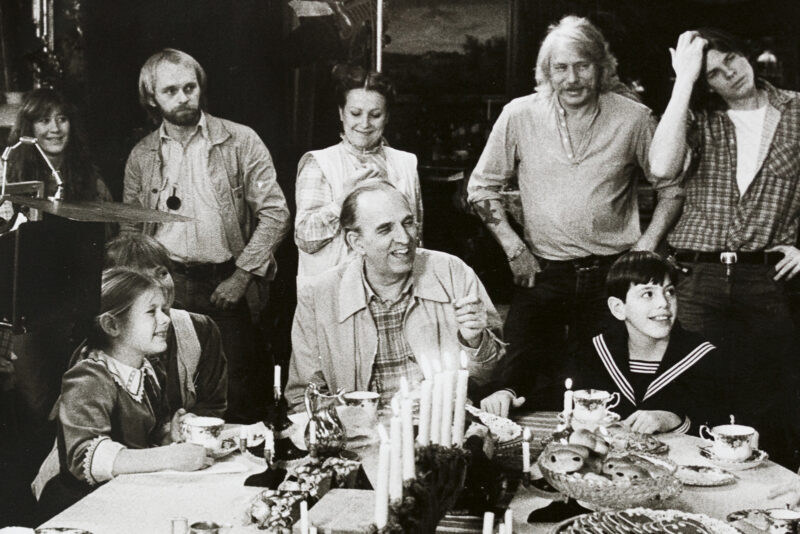 Ingmar Bergman sitter vid ett bord under inspelningen av Fanny och Alexander. Runtom honom sitter och står skådespelare och filmarbetare.