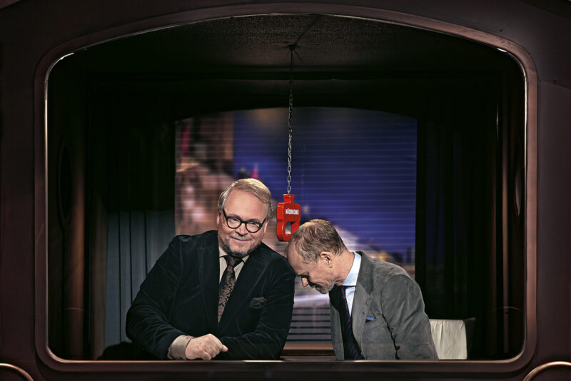 Bild på Fredrik Lindström och Kristian Luuk i en tågvagn i På spåret-studion. Kristian lutar sitt huvud mot Fredriks axel.